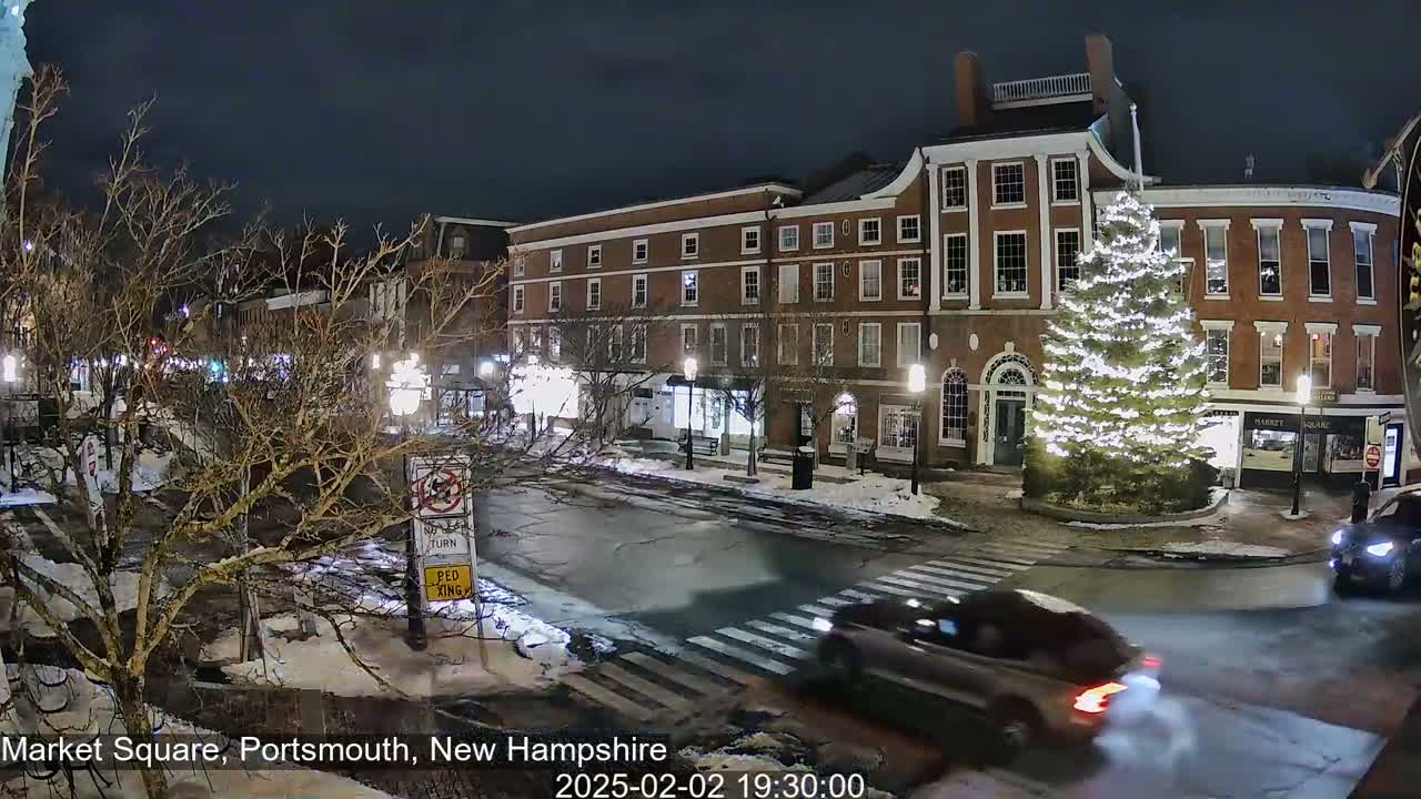 A snow-covered nighttime town square features a large, illuminated Christmas tree in front of brick buildings and several cars driving through the intersection.