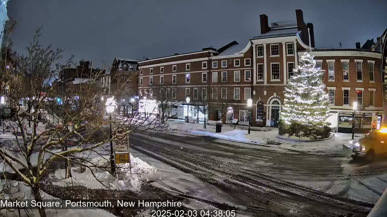 A snow-covered town square at night features a large illuminated Christmas tree in front of brick buildings, with a few cars visible on the road.