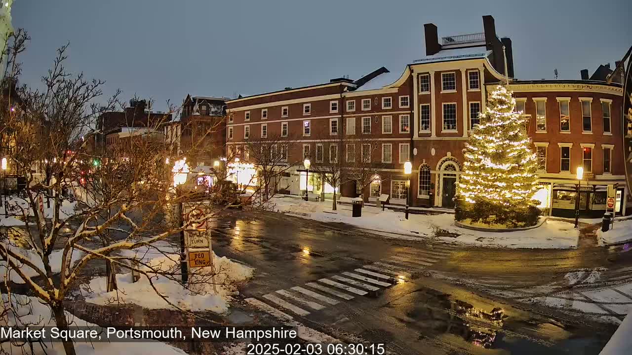 A snow-covered town square at night features a large, illuminated Christmas tree in front of brick buildings under a twilight sky.