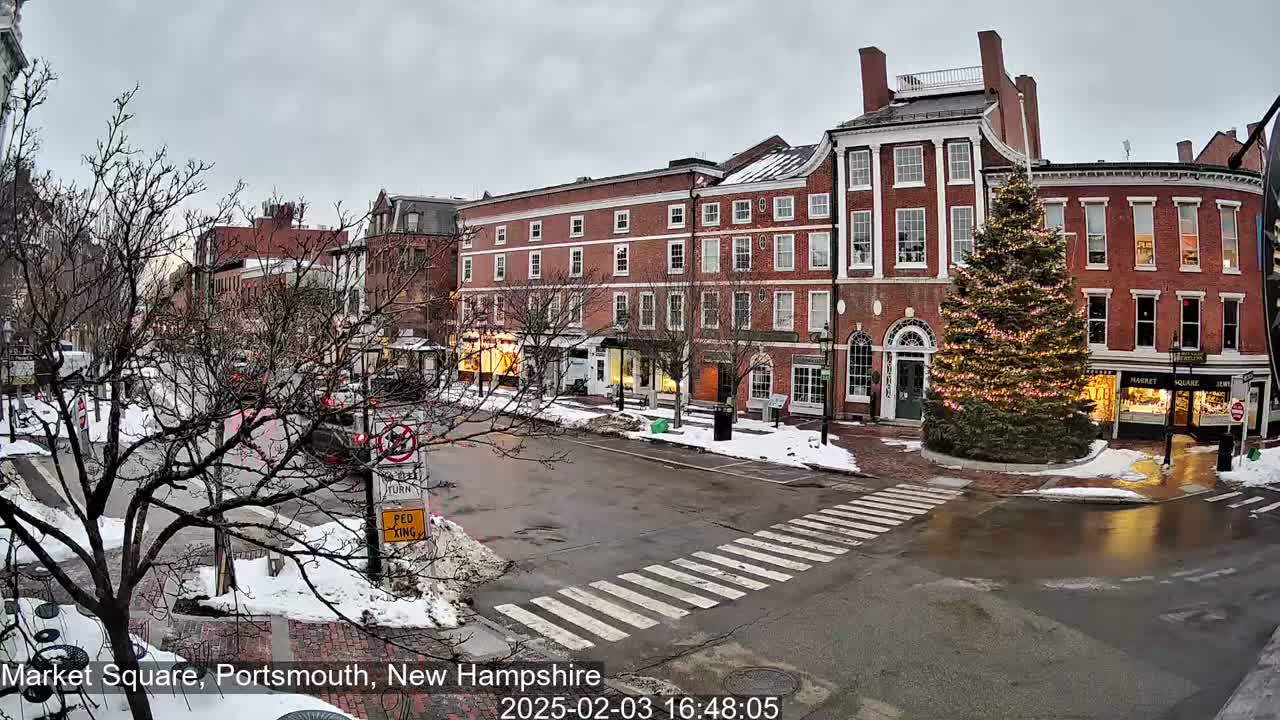 A snow-covered town square with brick buildings, a decorated Christmas tree, and a pedestrian crossing under an overcast sky.
