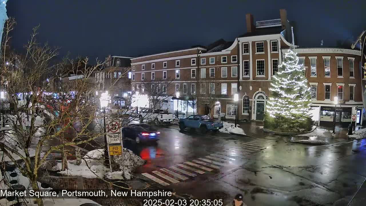 A nighttime view of a town square with brick buildings, a large illuminated Christmas tree, and wet streets with some cars and pedestrians.