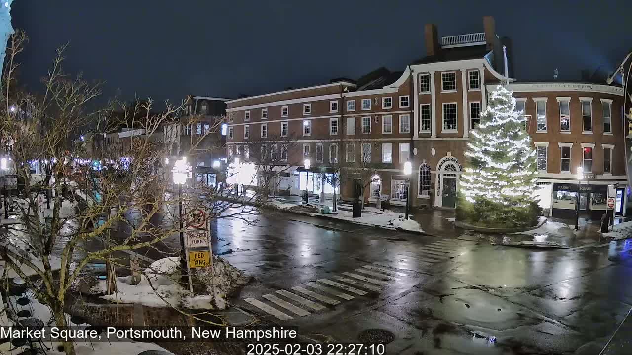 A nighttime view of a town square with brick buildings, a decorated Christmas tree, and wet streets under a dark sky.