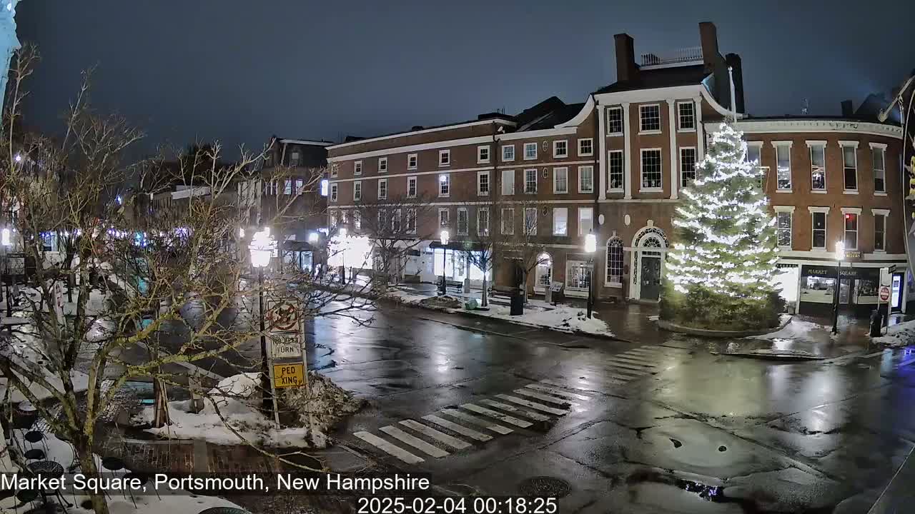 A wet, nighttime street scene shows a town square with brick buildings, a decorated Christmas tree, and patches of snow on the ground.