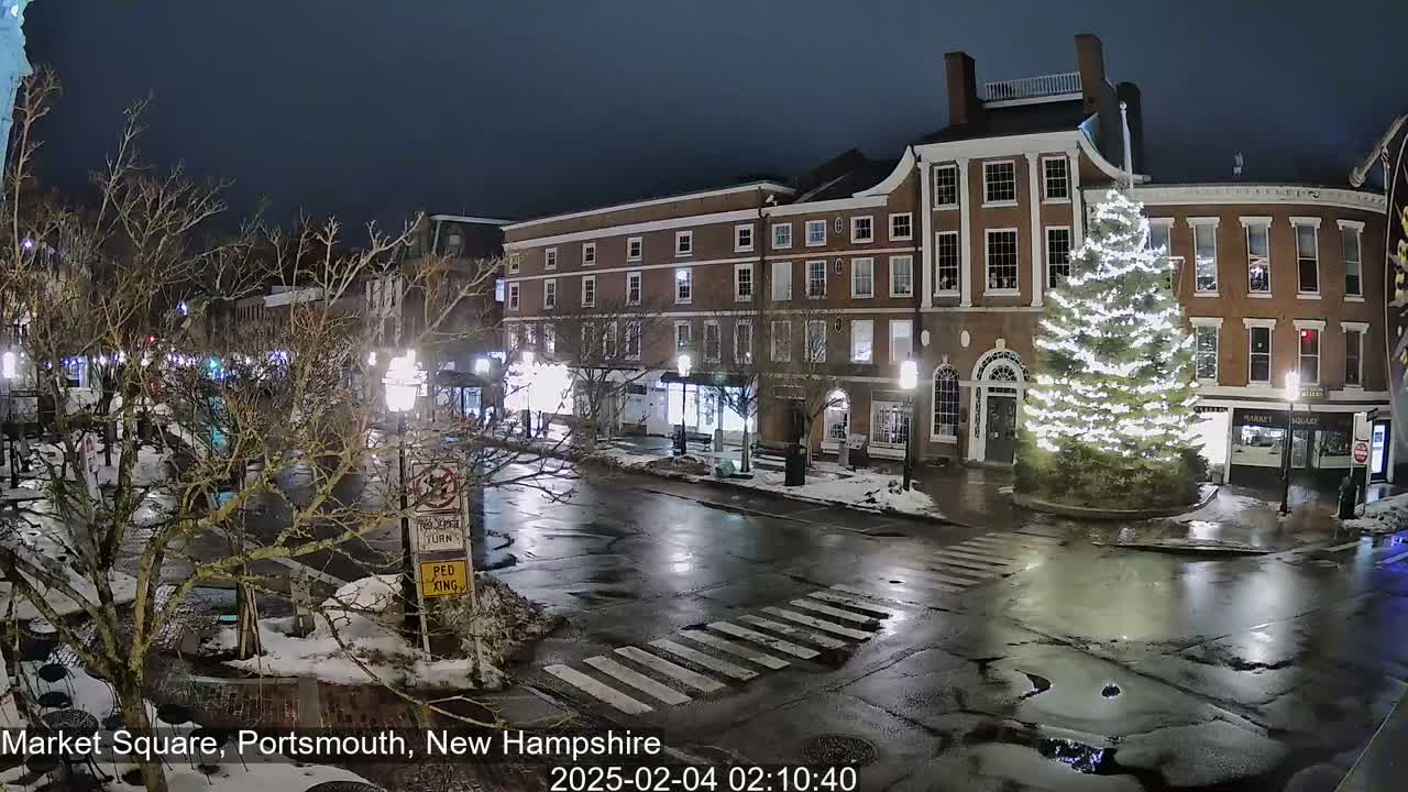 A wet, nighttime street scene shows a snow-dusted town square with brick buildings, a large illuminated Christmas tree, and bare trees.