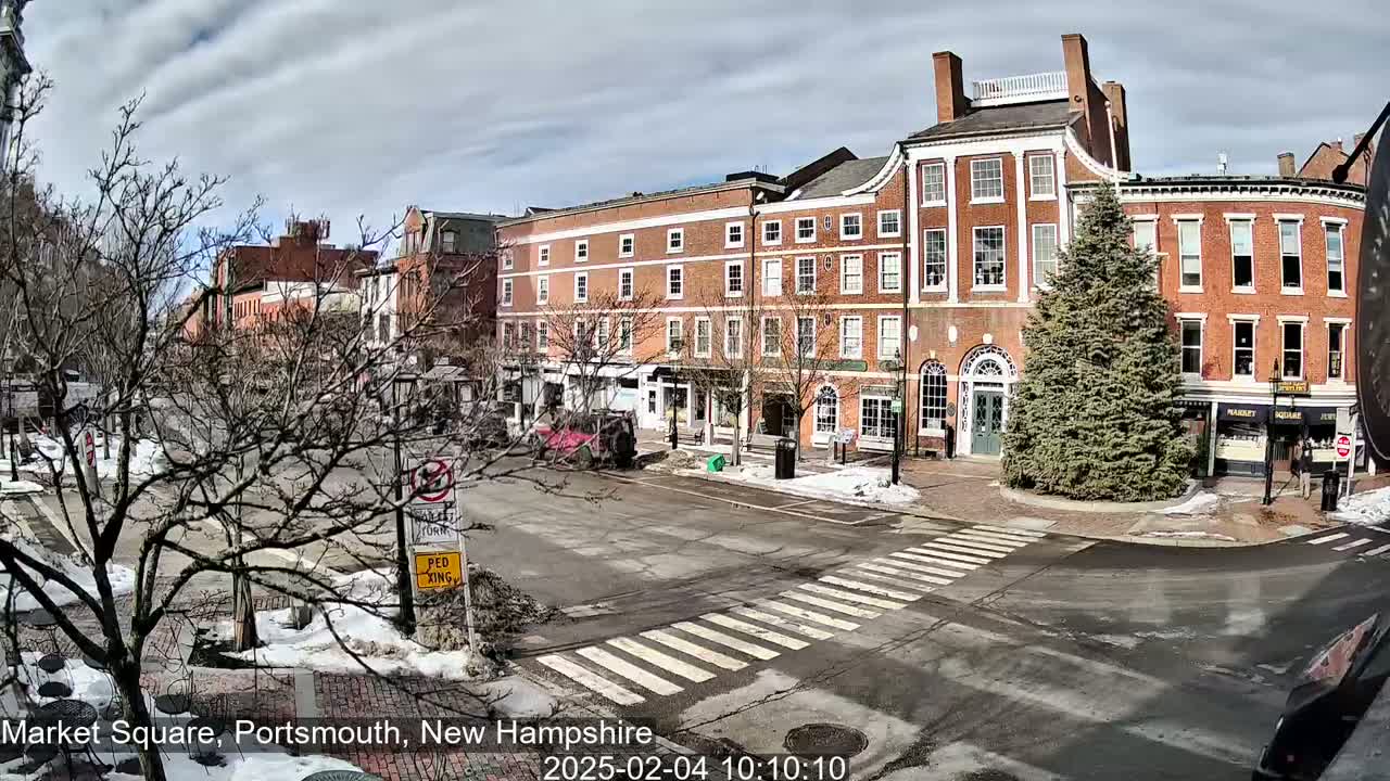 A partly sunny day reveals a town square with brick buildings, bare trees, and patches of snow and ice on the ground.