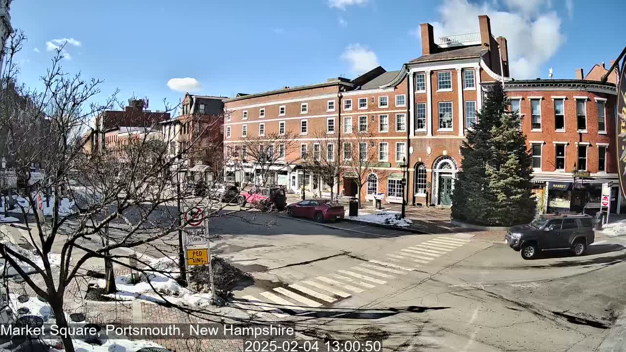 A sunny day in a brick-building-lined town square shows several cars parked and driving, with patches of snow and ice visible on the ground and sidewalks.