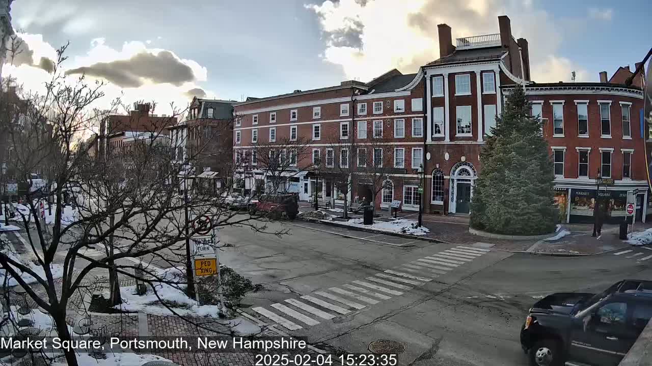 A partly cloudy day with some snow on the ground shows a brick building in a town square with cars parked and driving by.