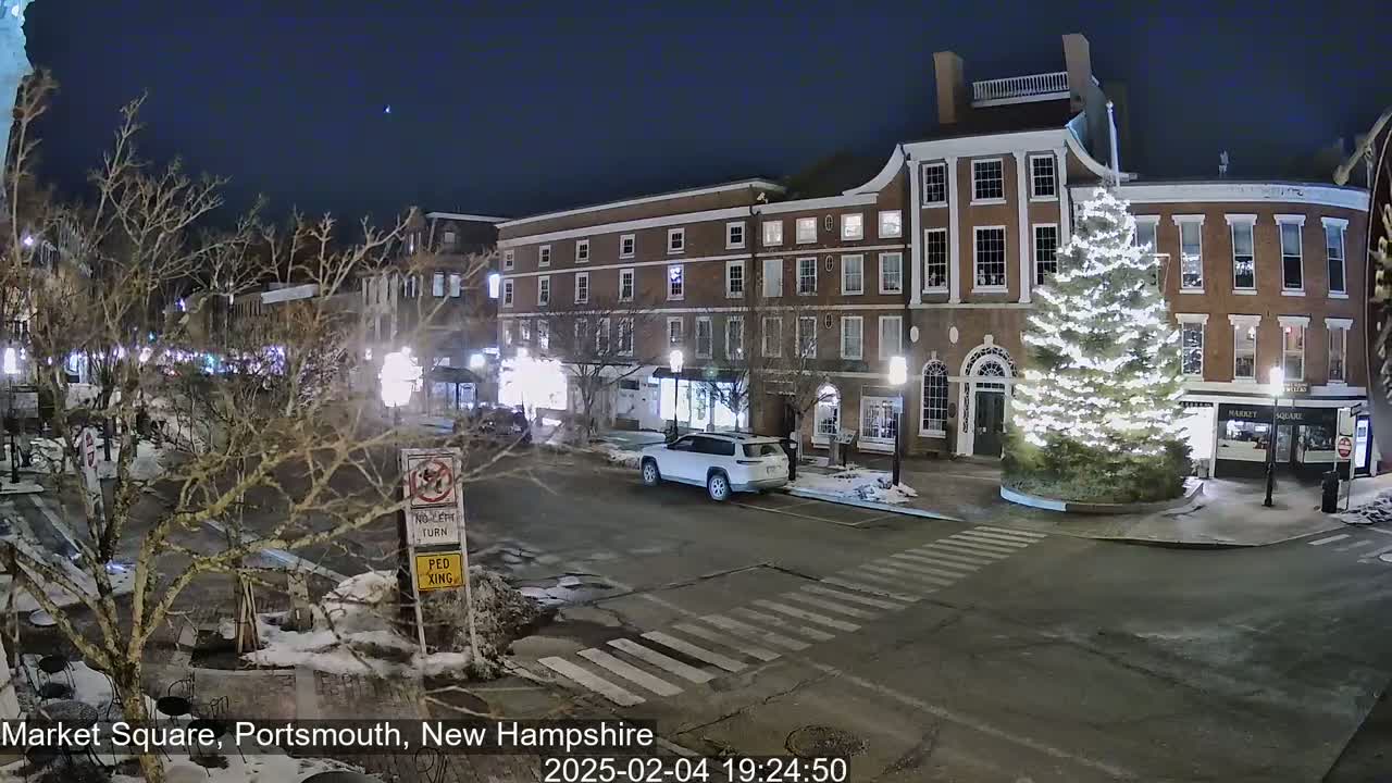 A snow-dusted night scene shows a town square with brick buildings, a large illuminated Christmas tree, and a single car parked on the street.