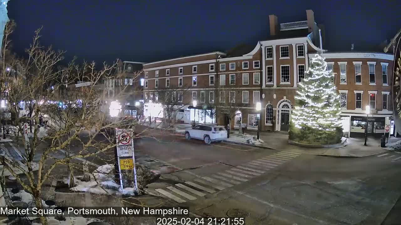 A snow-dusted town square at night, illuminated by streetlights, features a large, brightly lit Christmas tree in front of brick buildings.