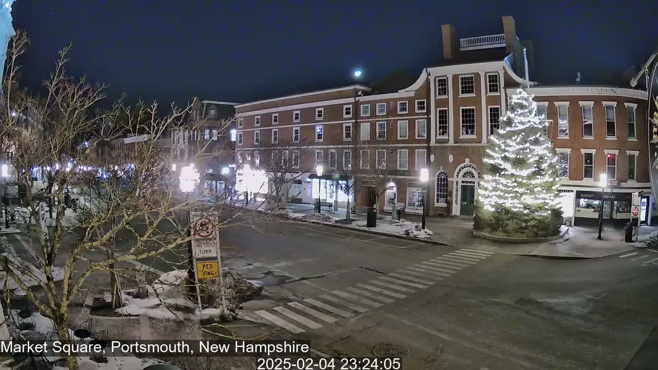 A snow-covered town square at night, illuminated by streetlights, features brick buildings, a large decorated Christmas tree, and sparsely-traveled roads.