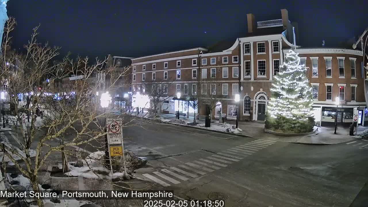 A snow-dusted, nighttime scene of a town square with brick buildings, a large illuminated Christmas tree, and bare trees lining a quiet street.