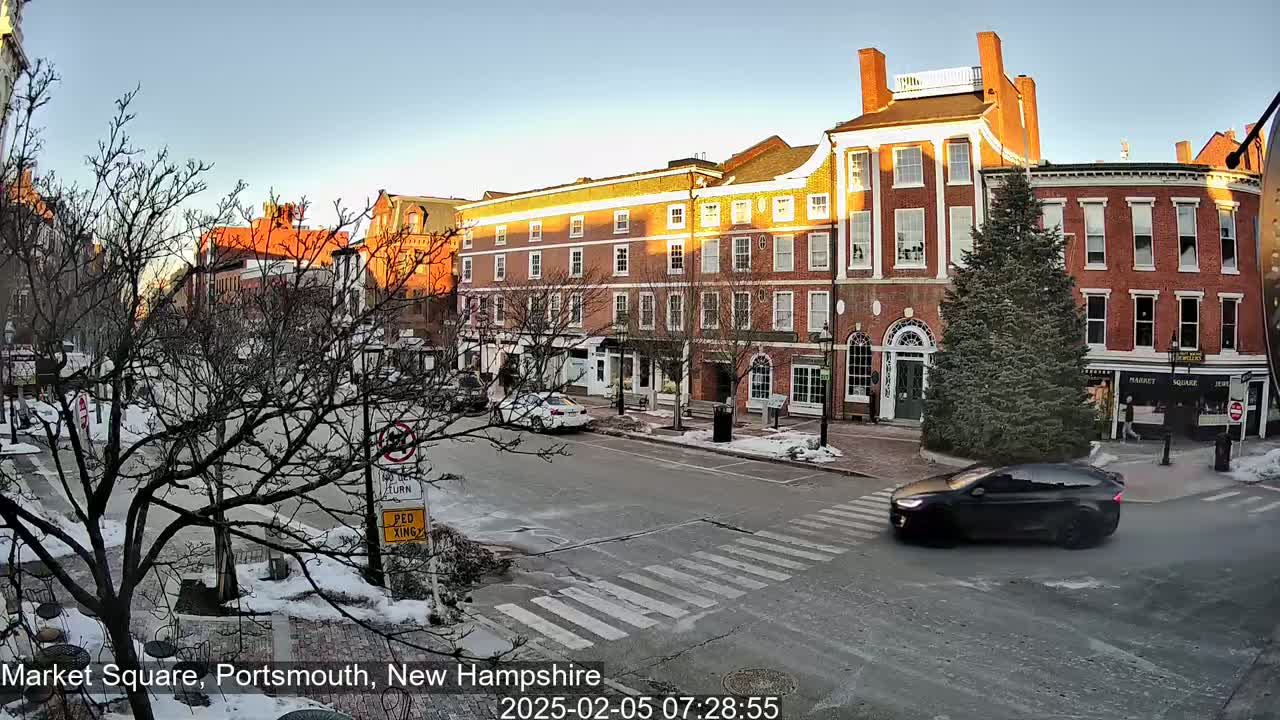 A dark-colored car drives across a snow-dusted street in front of brick buildings bathed in the golden light of a winter sunrise.