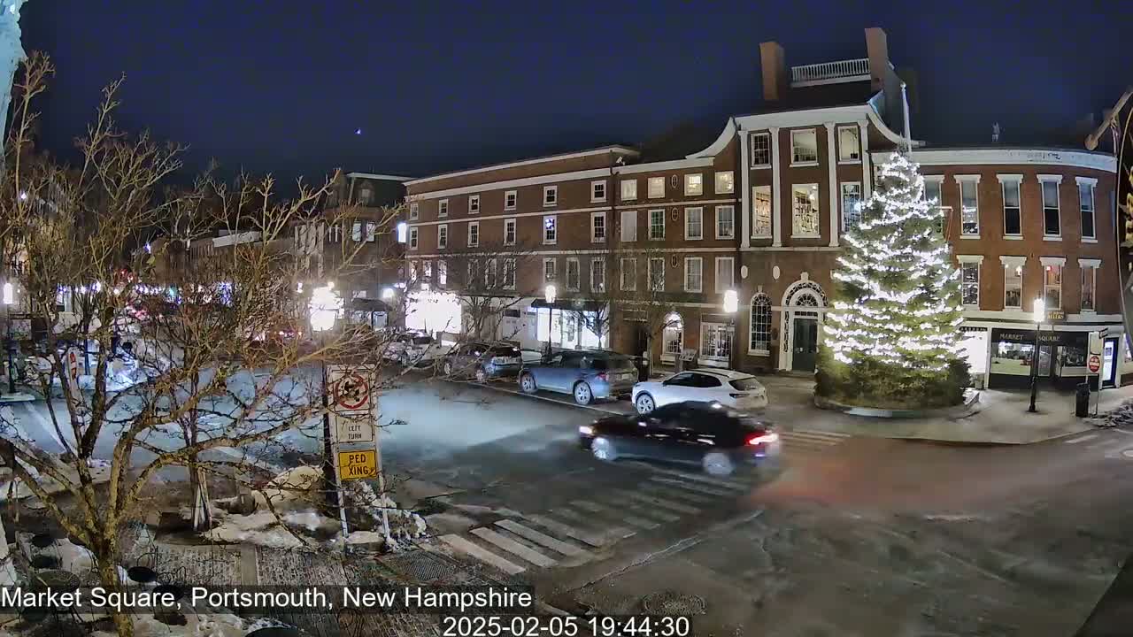 A nighttime scene of a snow-dusted town square with several cars driving past a large, illuminated Christmas tree in front of brick buildings.