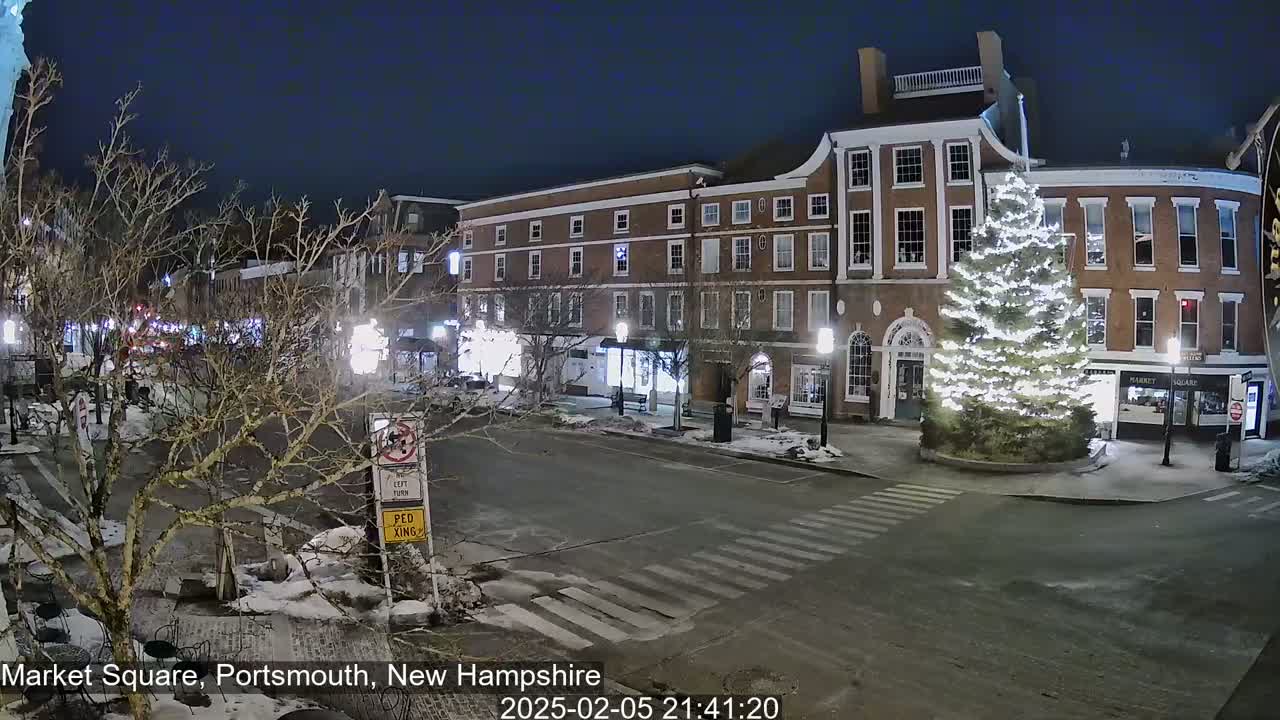 A snow-covered town square at night, illuminated by streetlights, features a large Christmas tree, brick buildings, and a crosswalk.