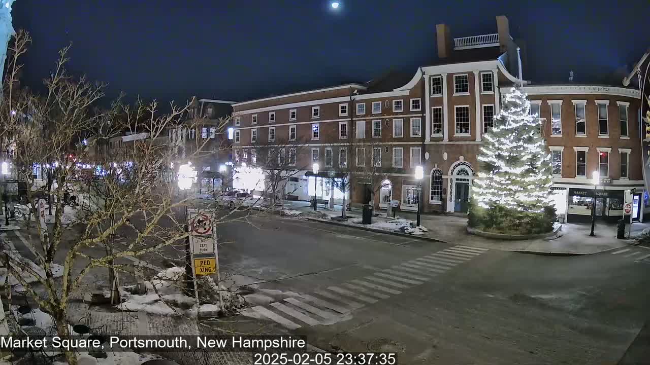 A snow-covered town square at night, illuminated by streetlights, features brick buildings and a large Christmas tree.