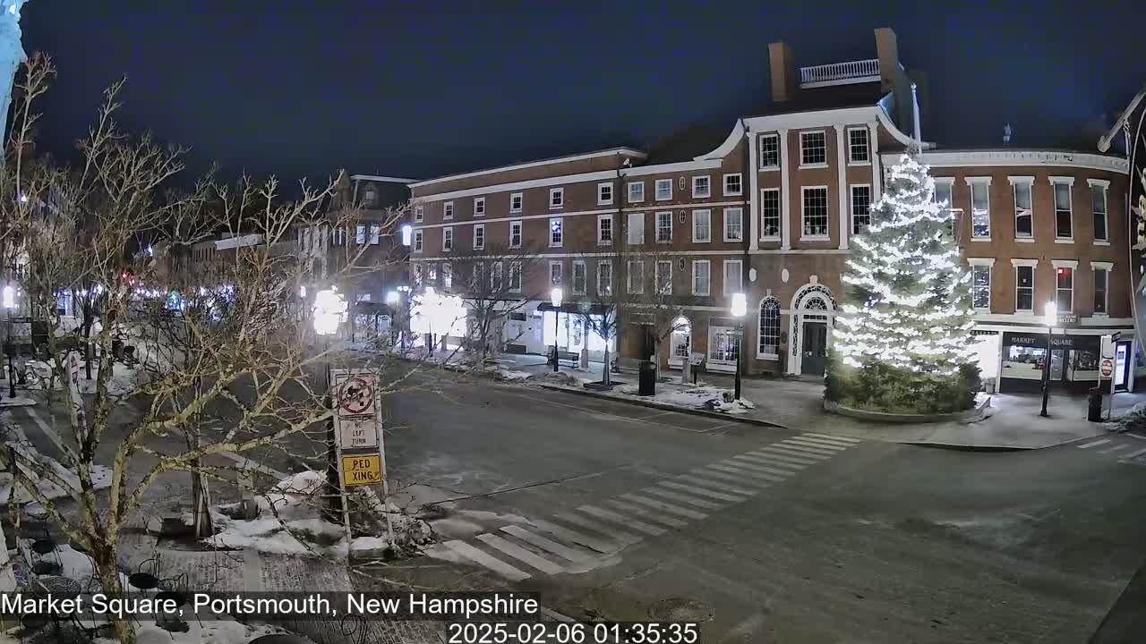 A snow-covered town square at night, illuminated by streetlights, features a large, decorated Christmas tree in the center and brick buildings lining the street.