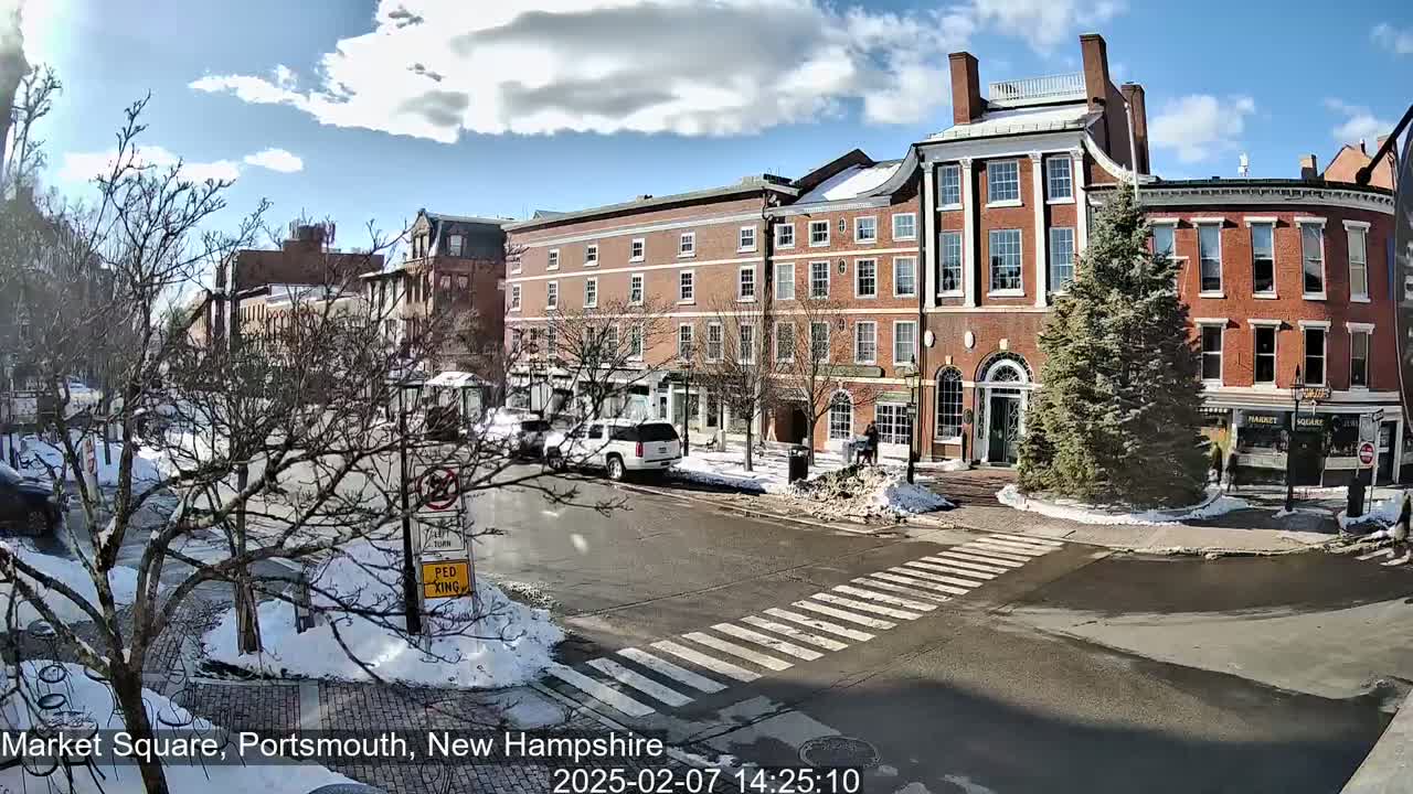 A partly sunny day reveals a brick-lined street scene with several multi-story buildings, a pedestrian crosswalk, parked cars, and snow patches on the ground.