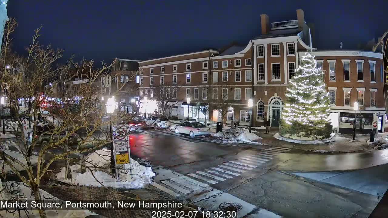A snow-dusted town square at night features a large, illuminated Christmas tree in front of brick buildings, with cars parked and driving on the street.