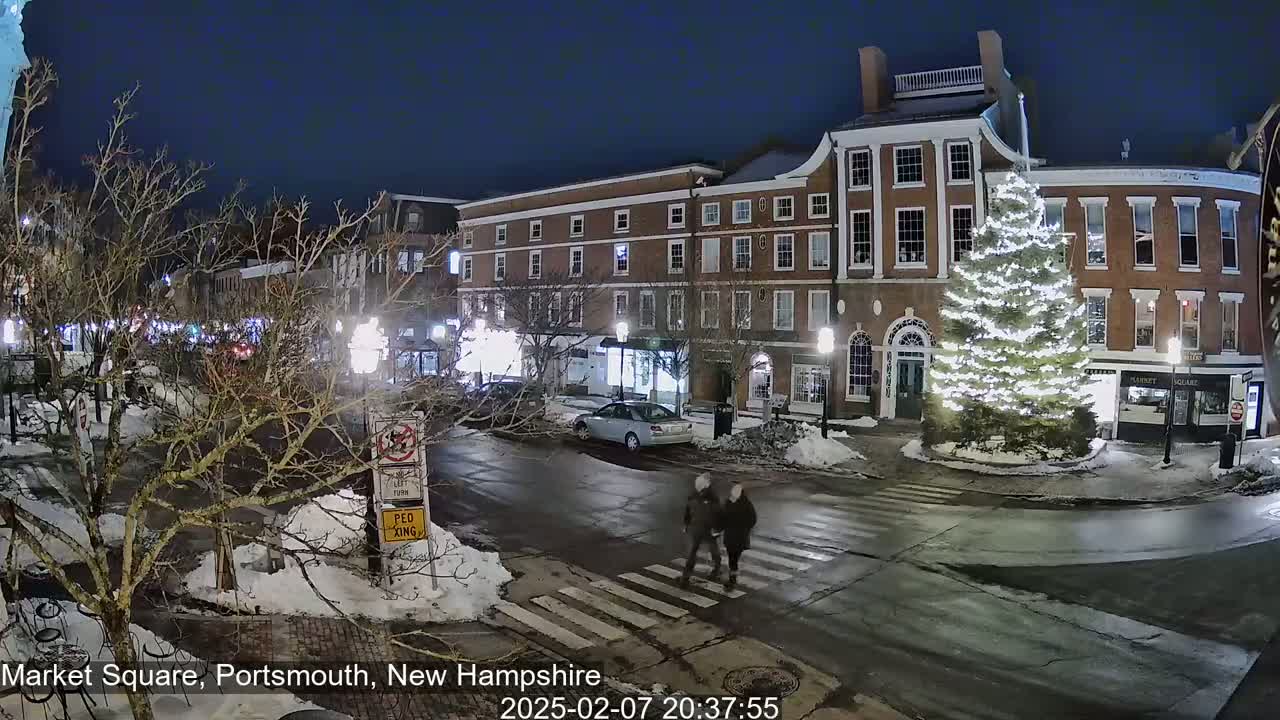 A nighttime view of a snow-covered town square with brick buildings, a large illuminated Christmas tree, and a couple crossing the street.