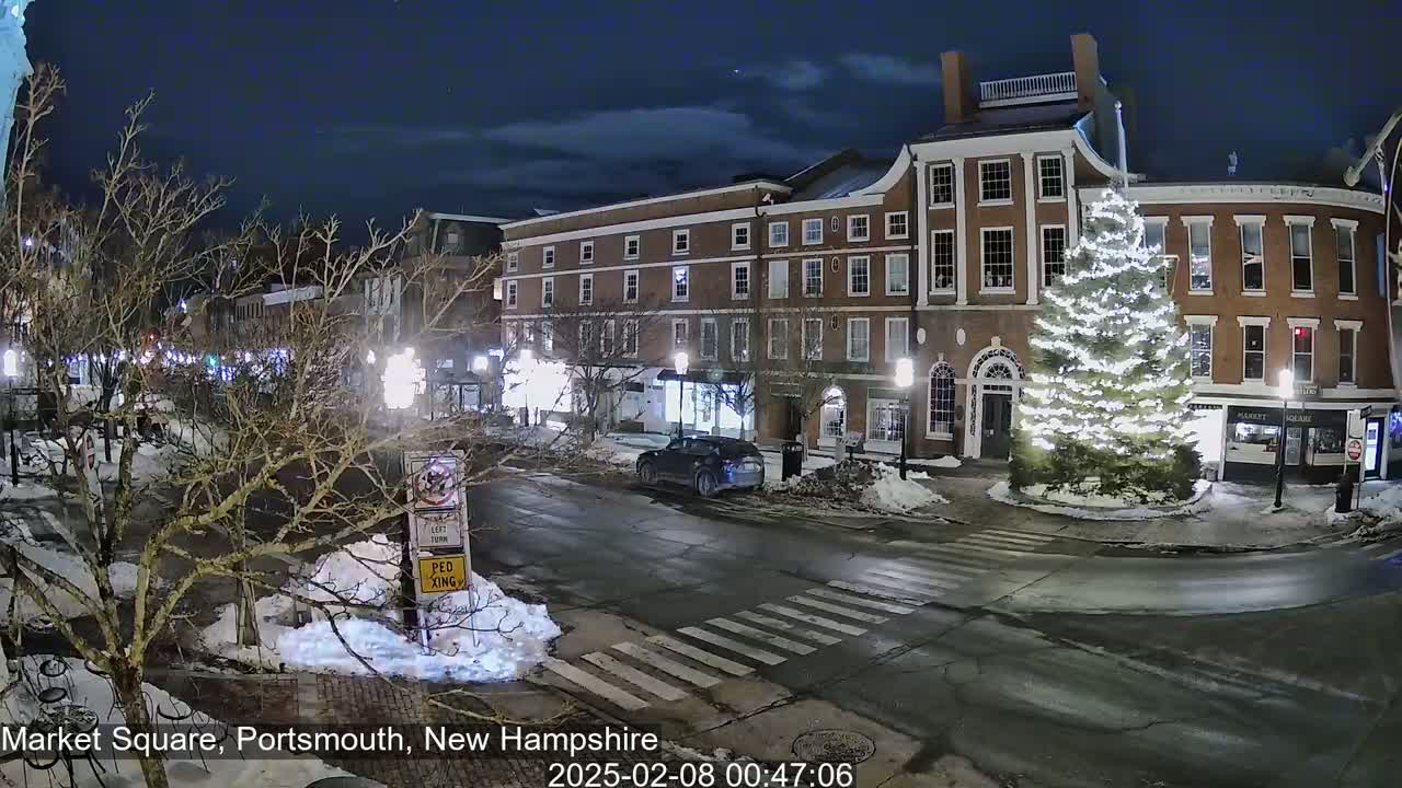 A nighttime street scene in a snow-covered town square shows brick buildings, a decorated Christmas tree, and a few cars under a partly cloudy sky.