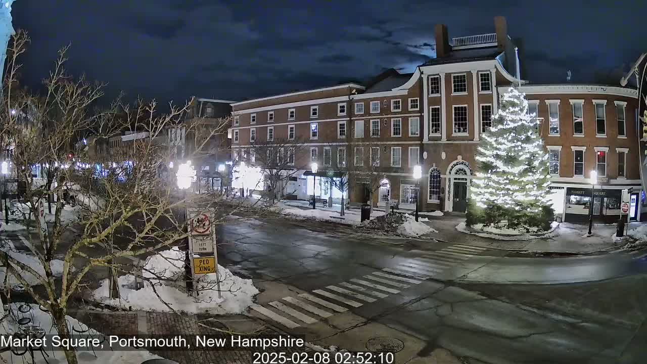 A snow-covered town square at night, illuminated by streetlights, features a large, decorated Christmas tree in the center, and brick buildings lining the streets.