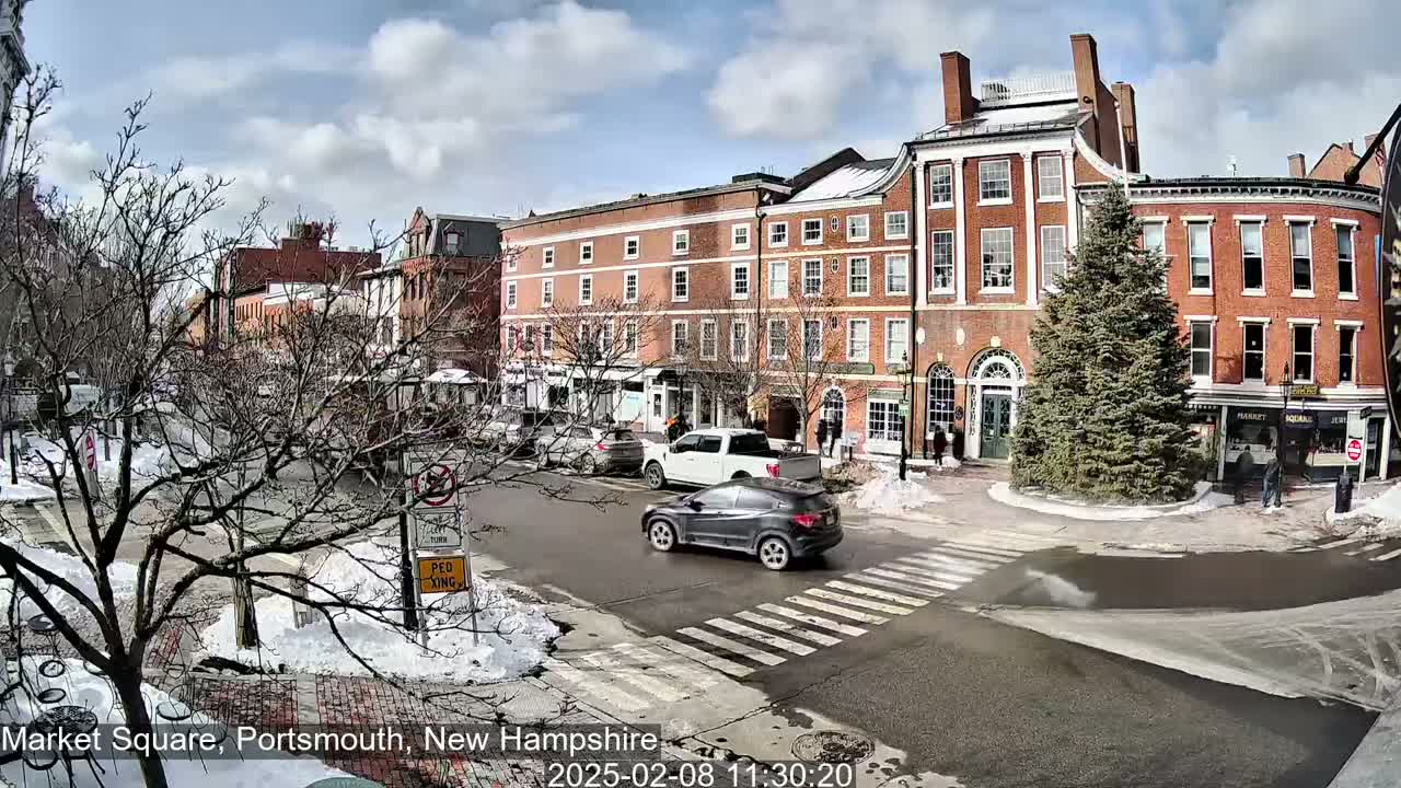 A sunny winter day in a snow-dusted town square shows several brick buildings, cars crossing an intersection, and a lone evergreen tree in the center.