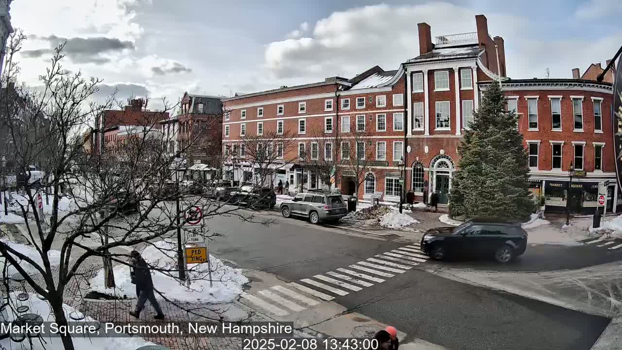 A partly cloudy day reveals a brick-lined town square with several cars and pedestrians, some snow and bare trees are visible.