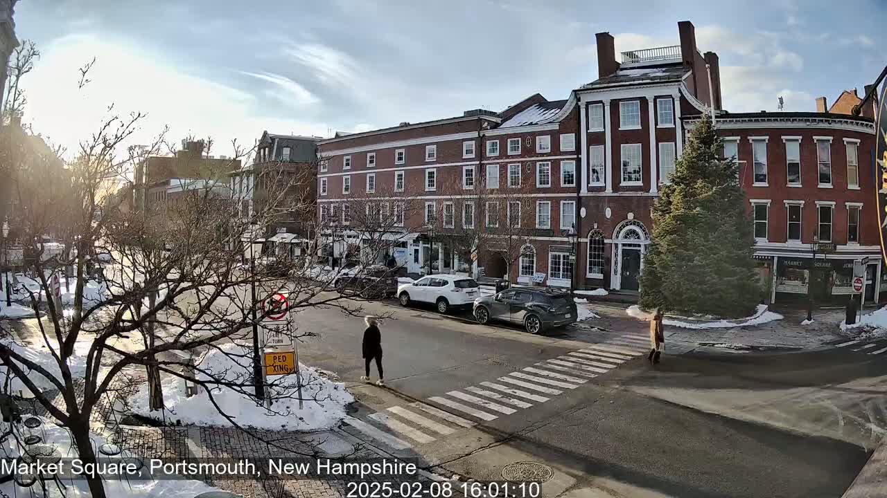 A person walks across a snow-dusted street in front of several brick buildings on a sunny day.