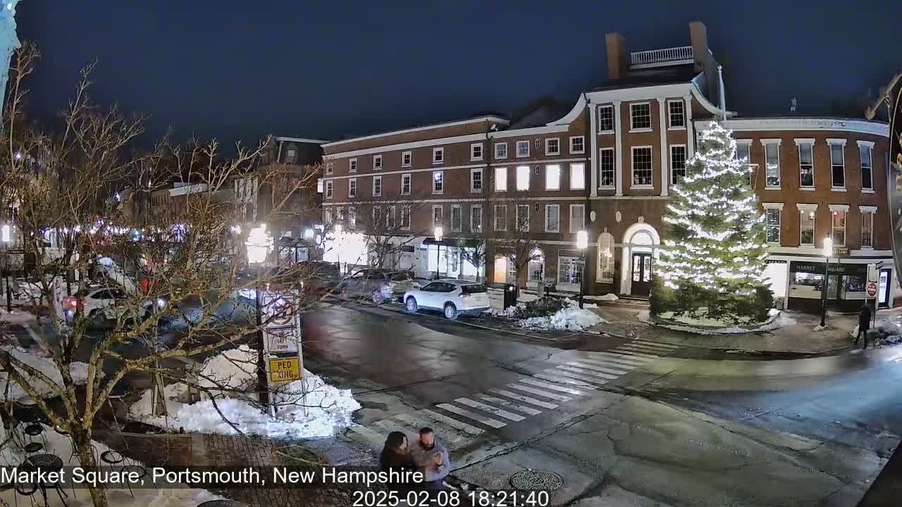 A snow-covered town square at night, illuminated by streetlights and a large, decorated Christmas tree, shows cars parked and driving around a pedestrian crossing.