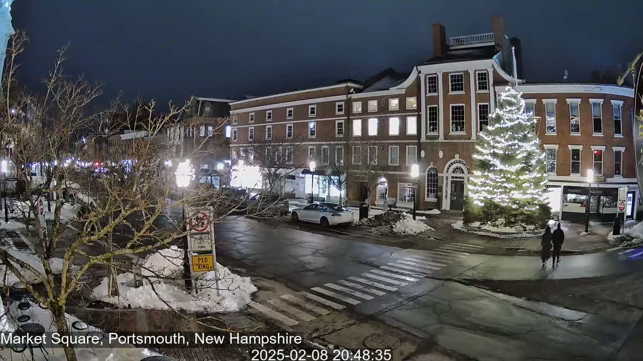 A snow-dusted town square at night features a large illuminated Christmas tree, brick buildings, parked cars, and pedestrians.