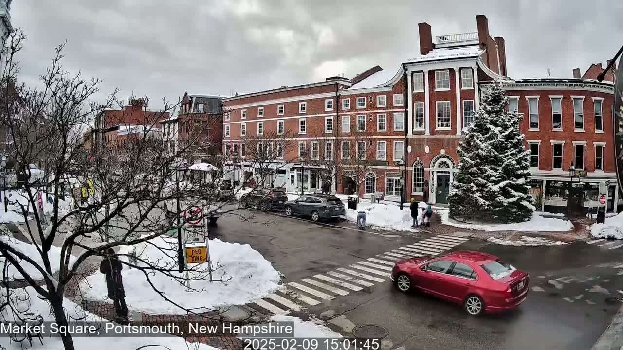 A snow-covered town square features several brick buildings, parked cars, pedestrians, and a red car driving through a crosswalk.