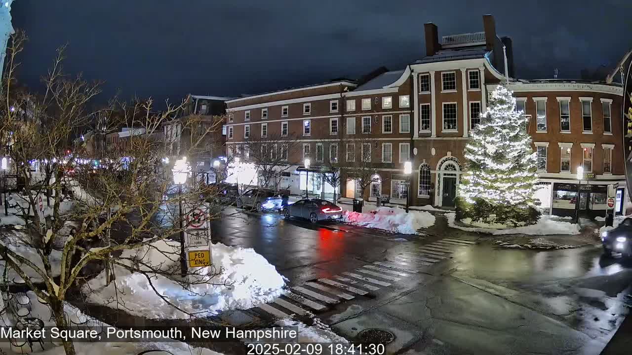 A snow-covered town square at night, illuminated by streetlights, features brick buildings, a large decorated Christmas tree, and a few cars driving through the intersection.