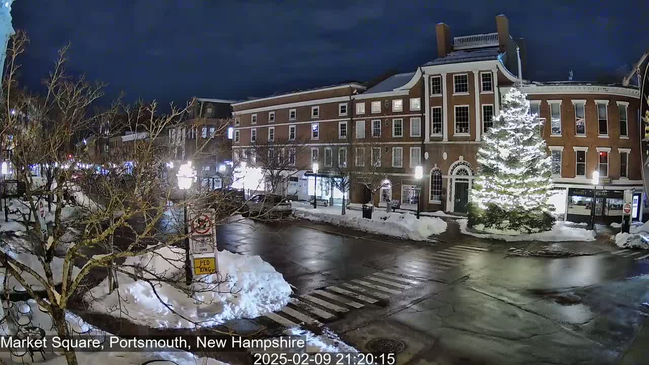 A snow-covered Market Square at night, illuminated by streetlights, features a large, brightly lit Christmas tree in the center and several brick buildings surrounding it.
