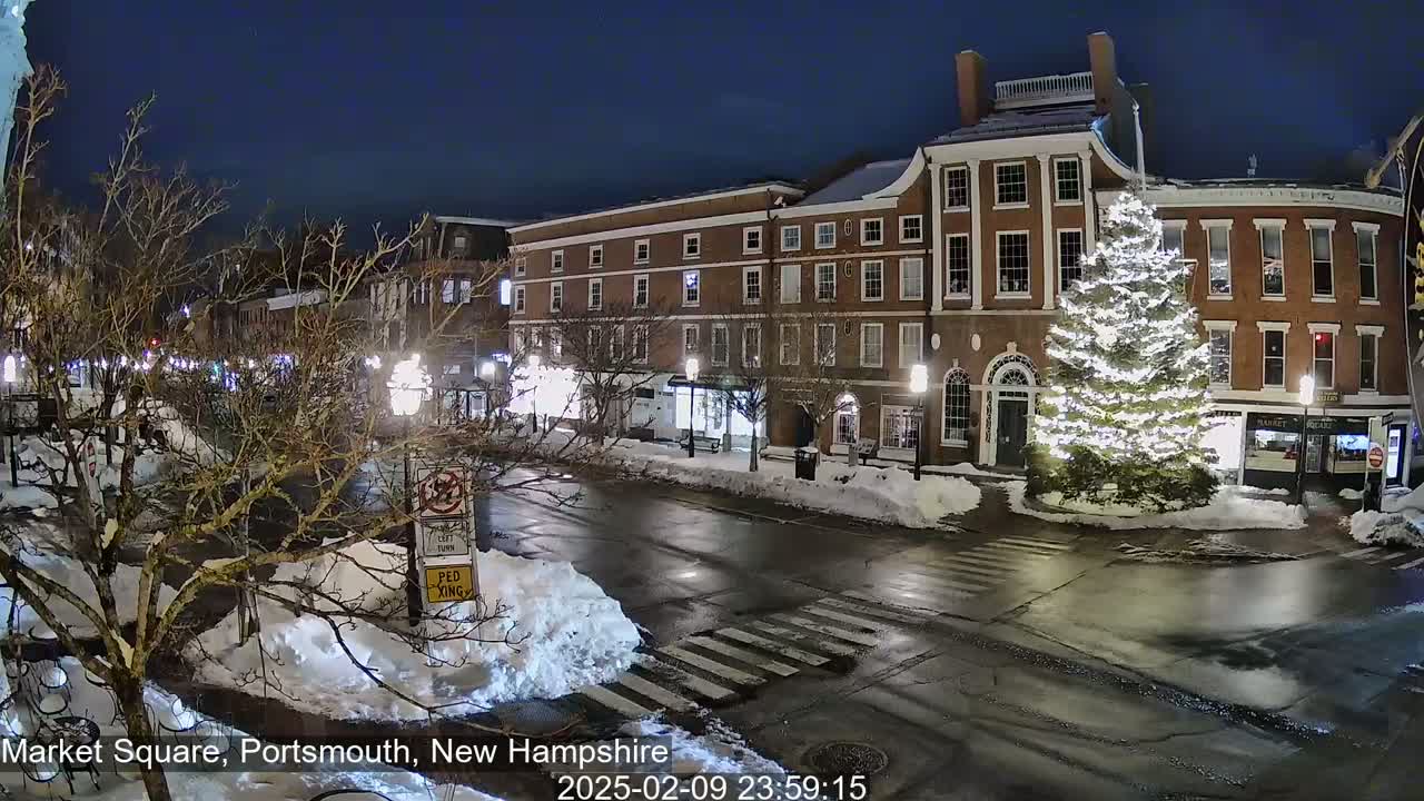 A snow-covered town square at night features a large, illuminated Christmas tree in front of brick buildings under a clear, dark sky.