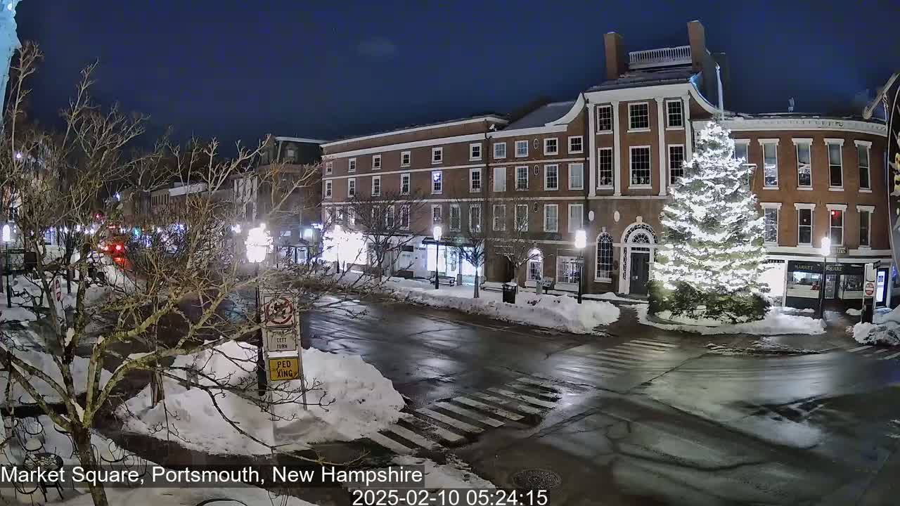 A snow-covered town square at night, illuminated by streetlights, features brick buildings, a large decorated Christmas tree, and a few cars on the road.