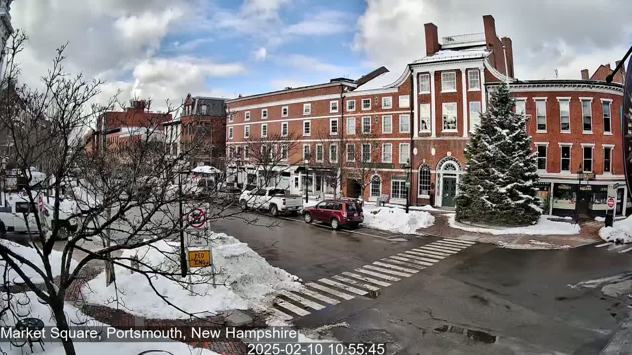 A snow-covered town square with brick buildings, parked cars, and a snow-covered evergreen tree in the center, under a partly cloudy sky.