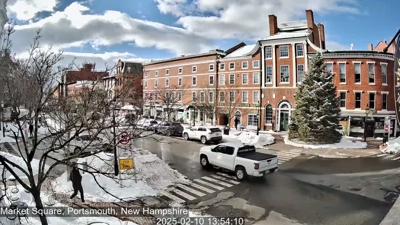 A snow-covered town square with brick buildings, cars, and pedestrians under a partly sunny sky.
