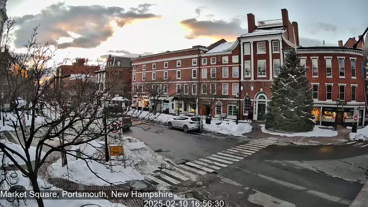 A snow-covered town square at sunset, with brick buildings, a few cars, and a large evergreen tree in the center.