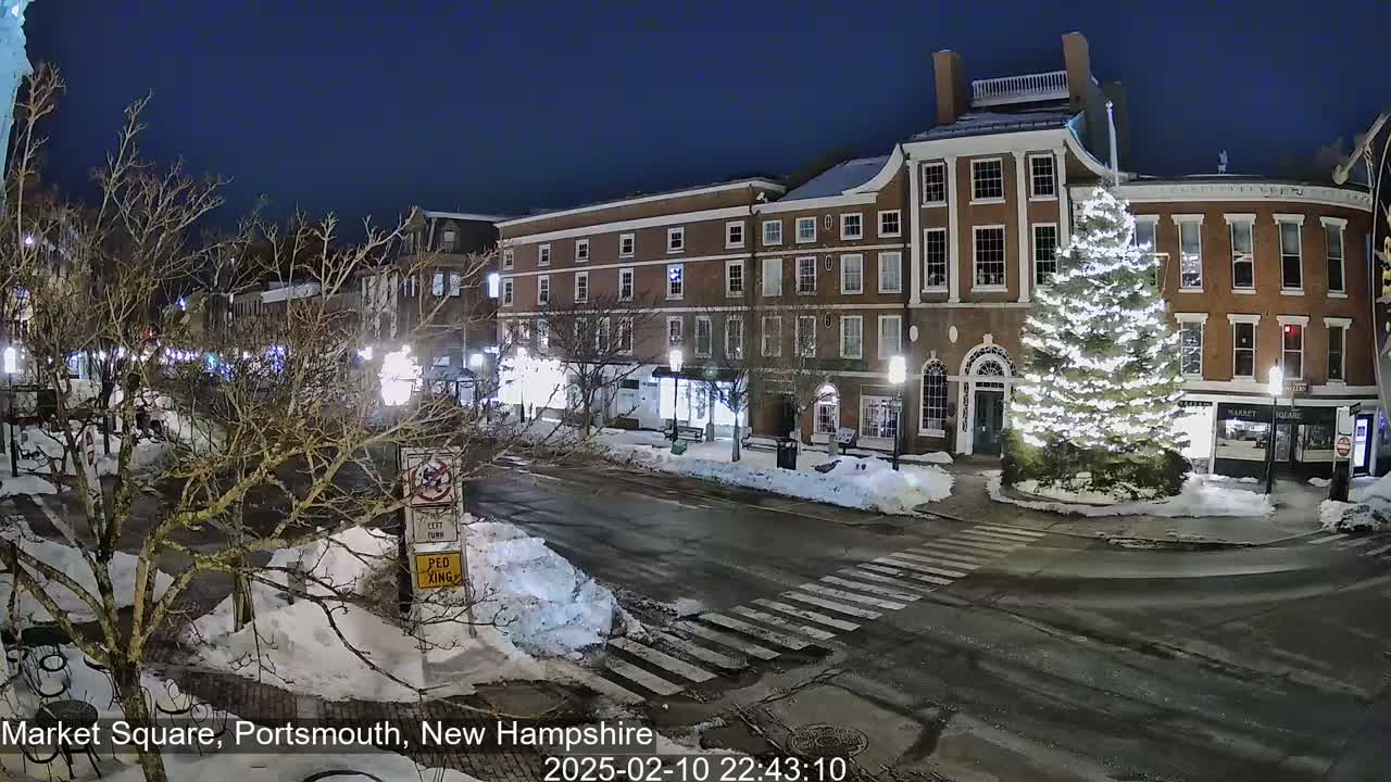 A snow-covered town square at night, featuring a large illuminated Christmas tree in front of brick buildings.