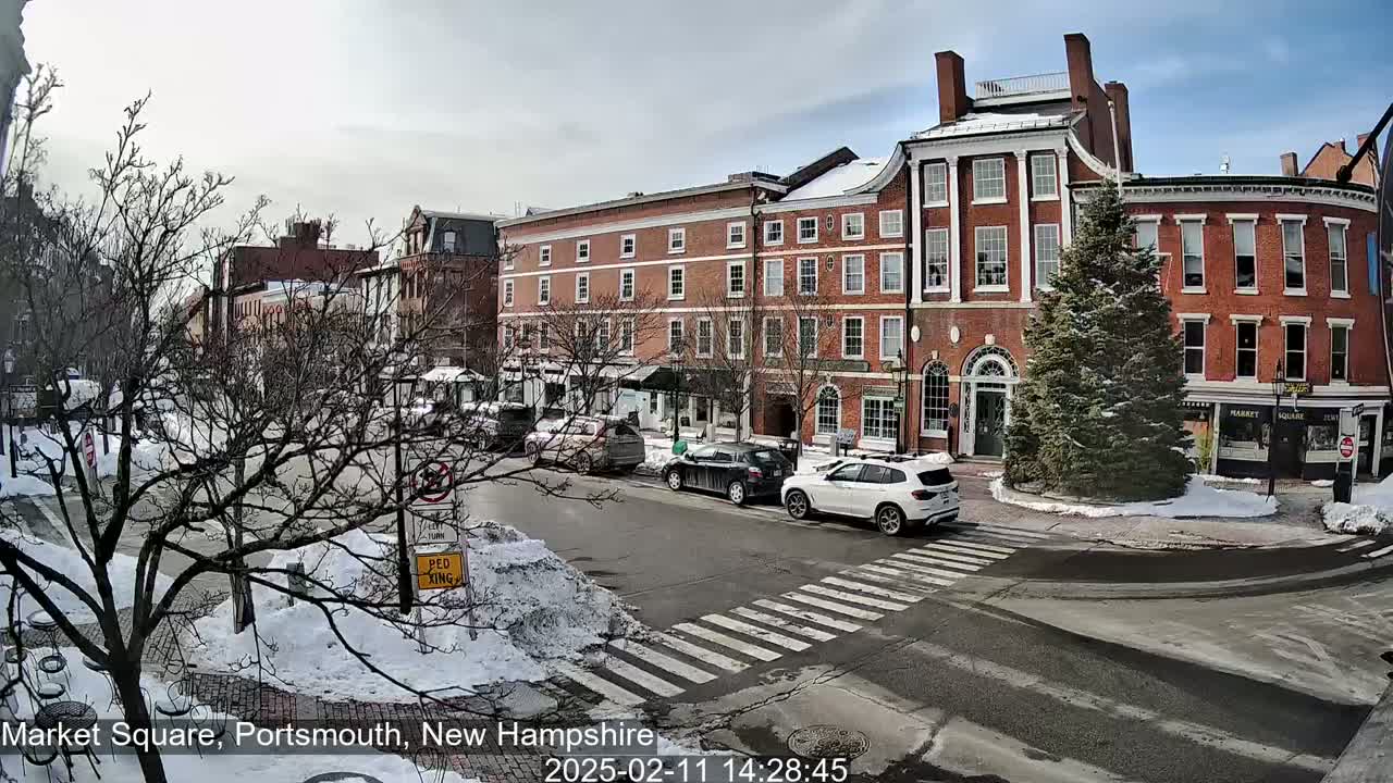 A snow-covered town square features several brick buildings, a few cars, and a pedestrian crossing under a sunny sky.