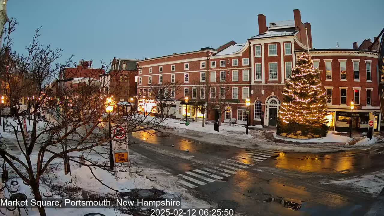 A snow-covered town square at twilight, featuring illuminated storefronts, a large decorated Christmas tree, and wet roads reflecting streetlights.