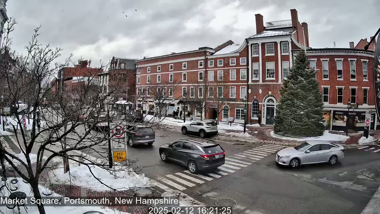 A snow-covered city square with several brick buildings, cars at an intersection, and a few pedestrians under an overcast sky.