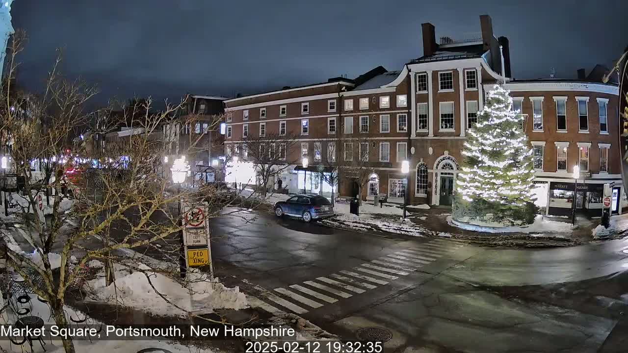 A snow-covered town square at night features a large illuminated Christmas tree in front of brick buildings, with a few cars and pedestrians visible under a dark sky.