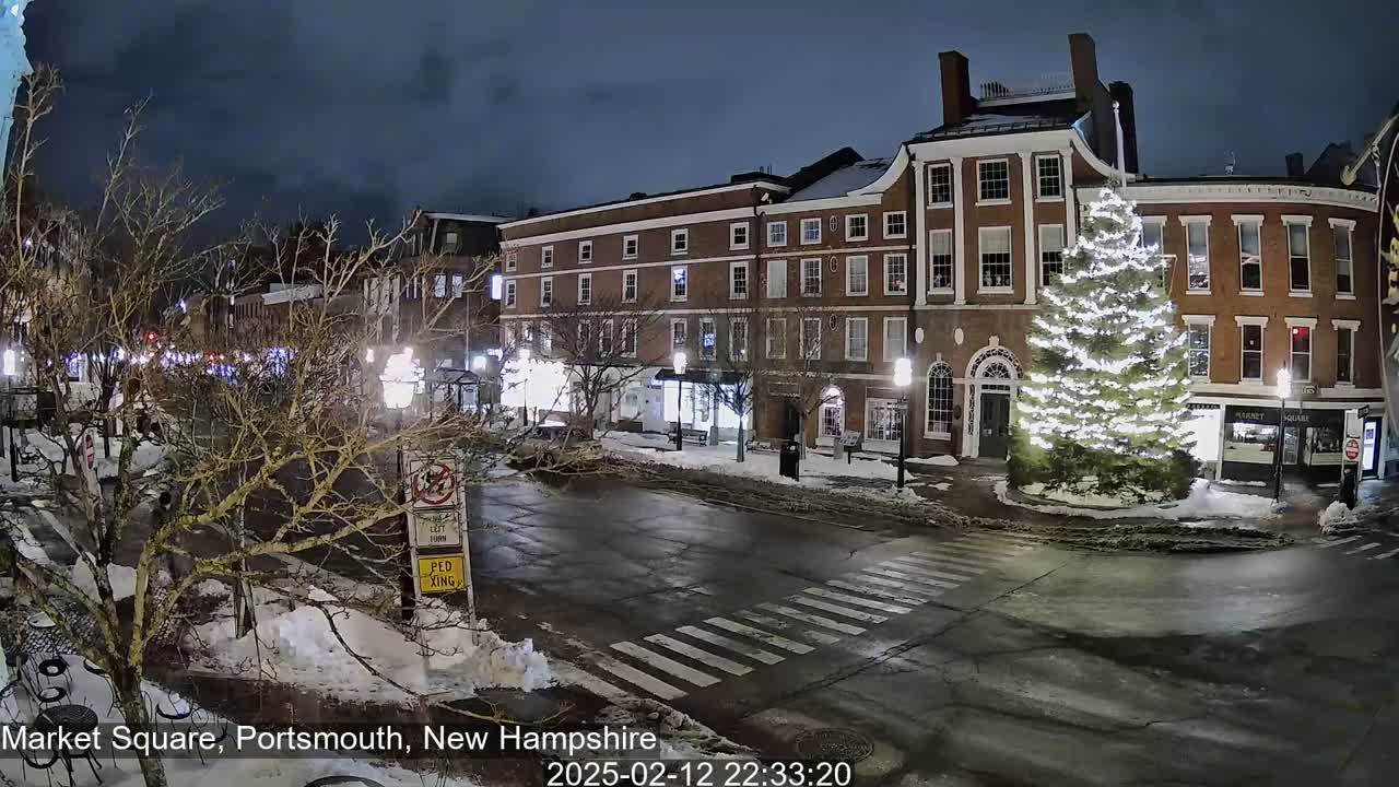 A snow-covered, nighttime street scene shows a large illuminated Christmas tree in a town square, flanked by brick buildings.