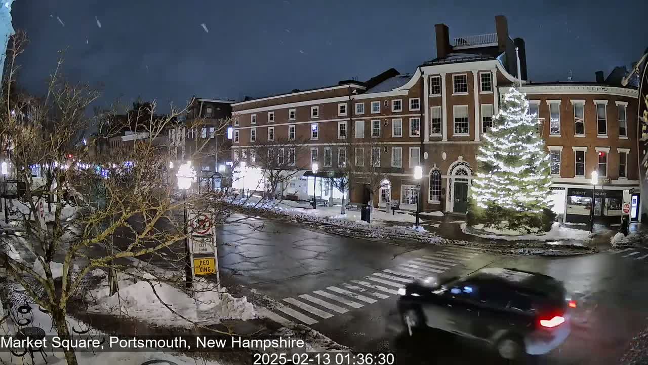 A snow-covered town square at night, featuring a large illuminated Christmas tree and a car driving through a crosswalk, under a dark, slightly cloudy sky.
