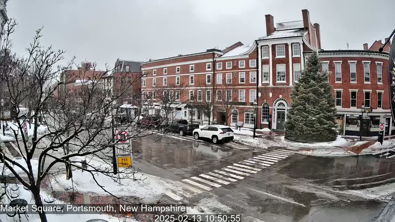A snow-covered town square with several brick buildings, bare trees, and a few cars parked along the street.
