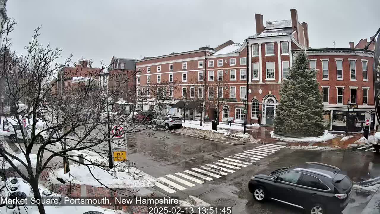 A snow-covered town square with brick buildings, bare trees, and a dark-colored SUV driving across a pedestrian crossing on a cloudy day.