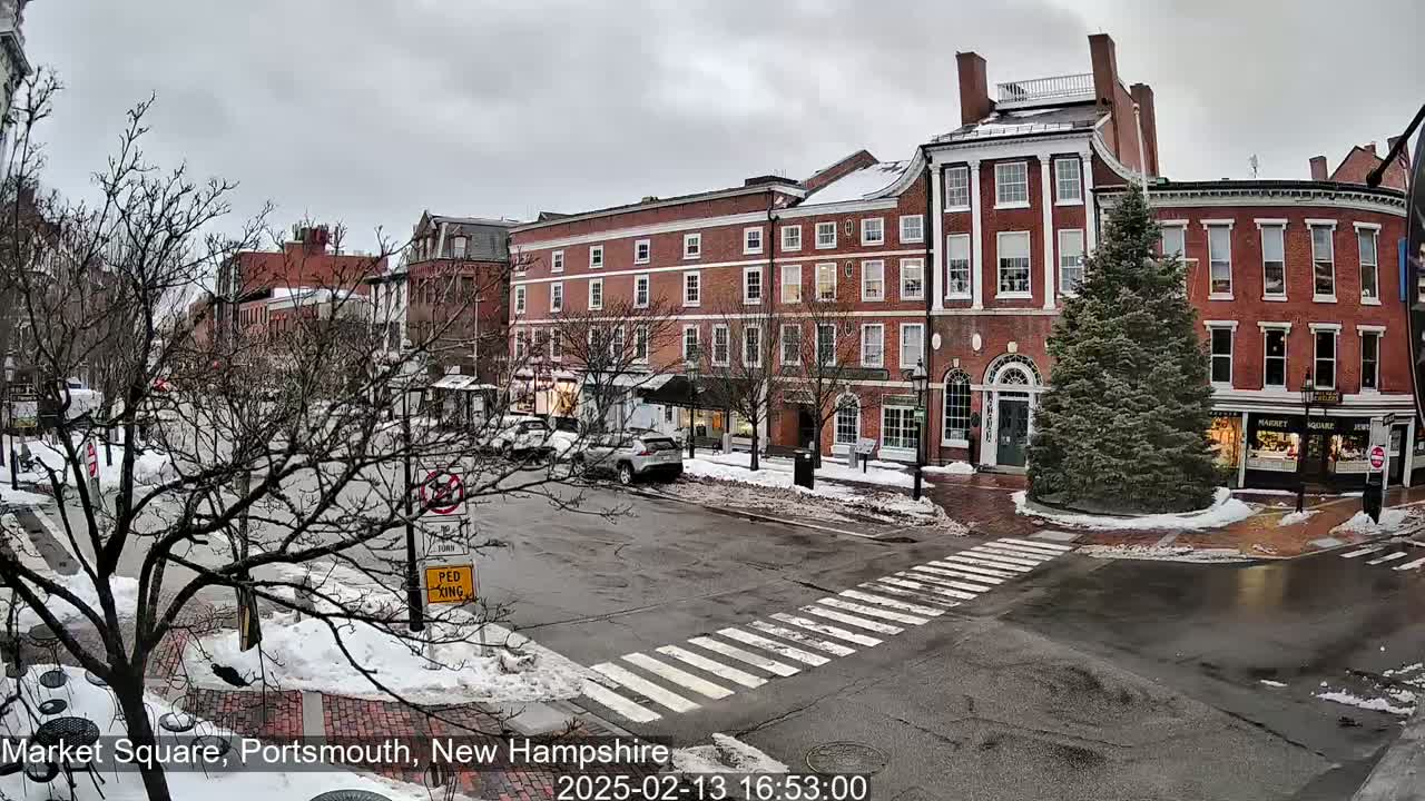 A snow-covered street scene shows a city square with several brick buildings and a few cars parked along the side of the road under an overcast sky.