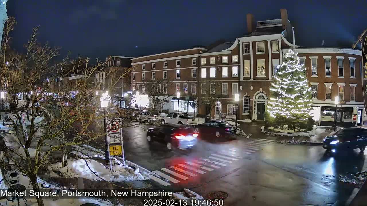 A nighttime view of a town square with several brick buildings, a large illuminated Christmas tree, and cars driving on wet roads with some snow on the ground.