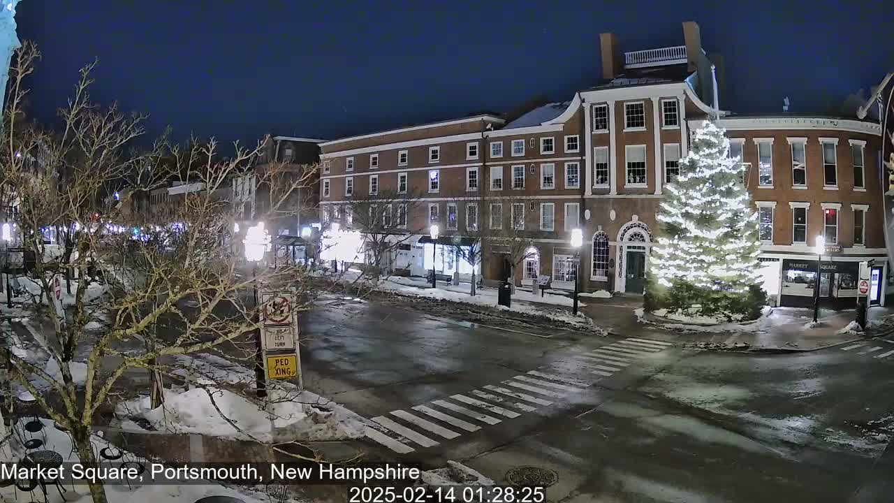 A snow-covered town square at night, illuminated by streetlights, features brick buildings and a large, lit Christmas tree.